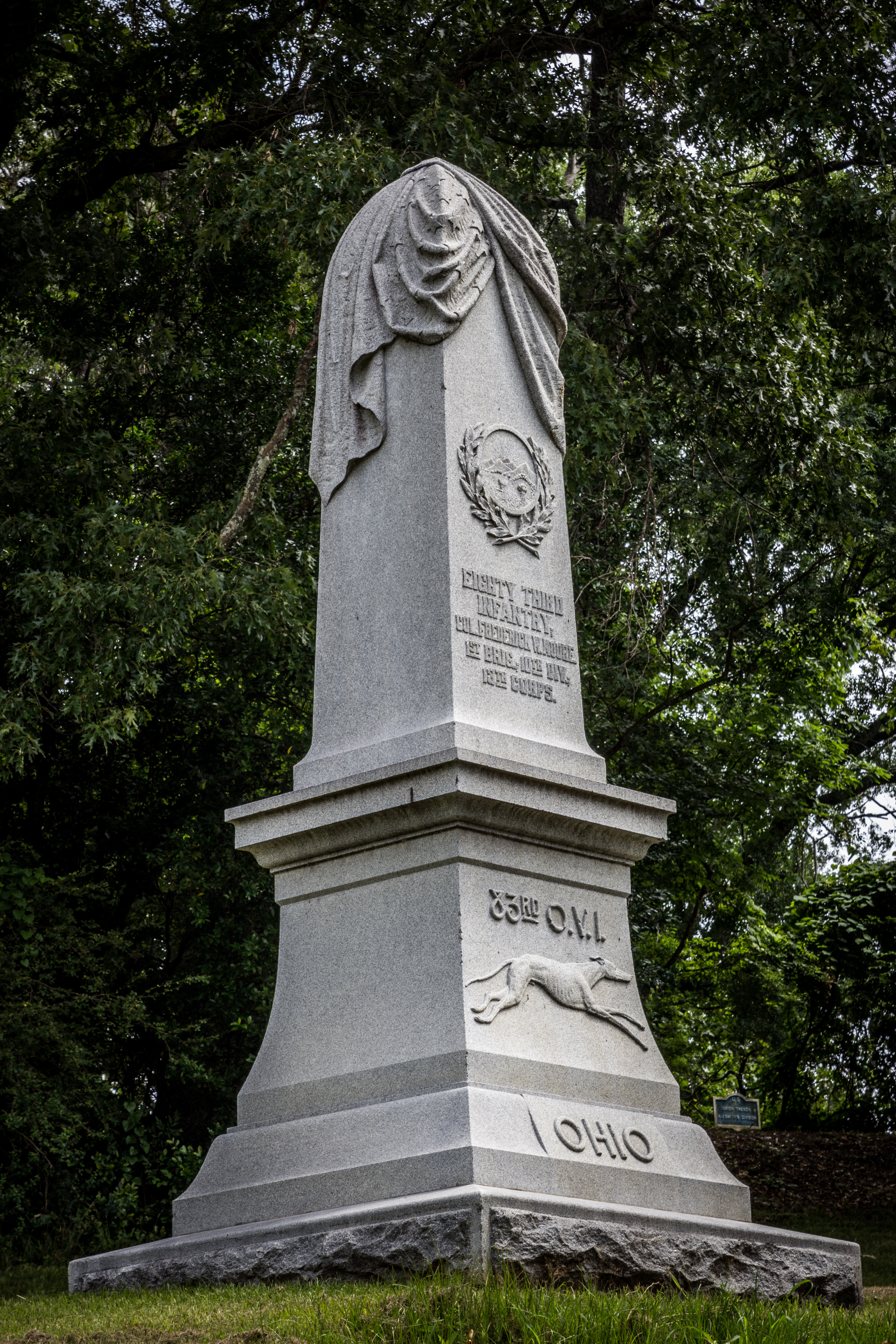 A tall vertical statue sits on a hill with a short inscription on the front of it
