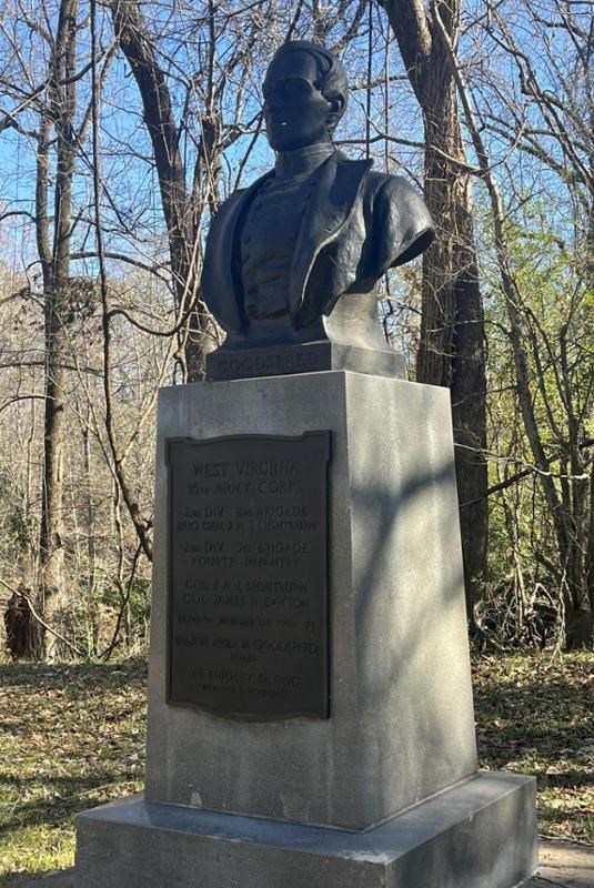 bronze bust of man in military uniform set on top of a stone pedestal, set against a wooded background with a blue sky. The base of the bust says "Goodspeed".