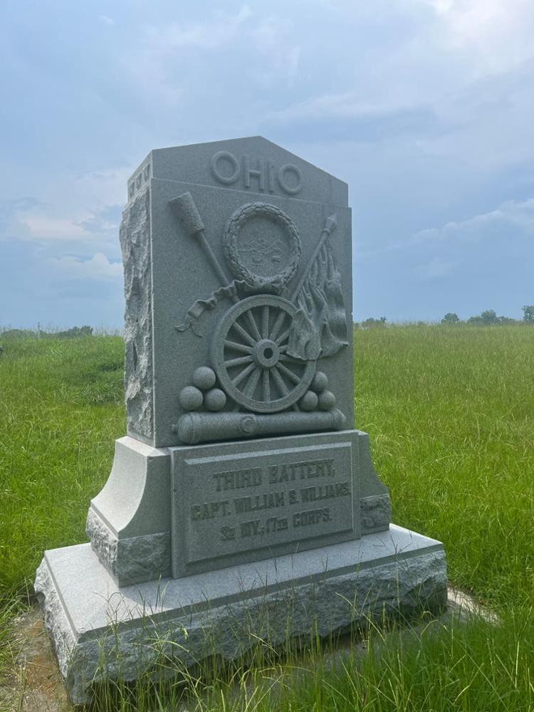 Stone monument with a pointed top, A spoked wheel in the center on top of a cannon and cannonballs. A sponge rammer rises at an angle on the left side, while an American rises at an angle on the right side.