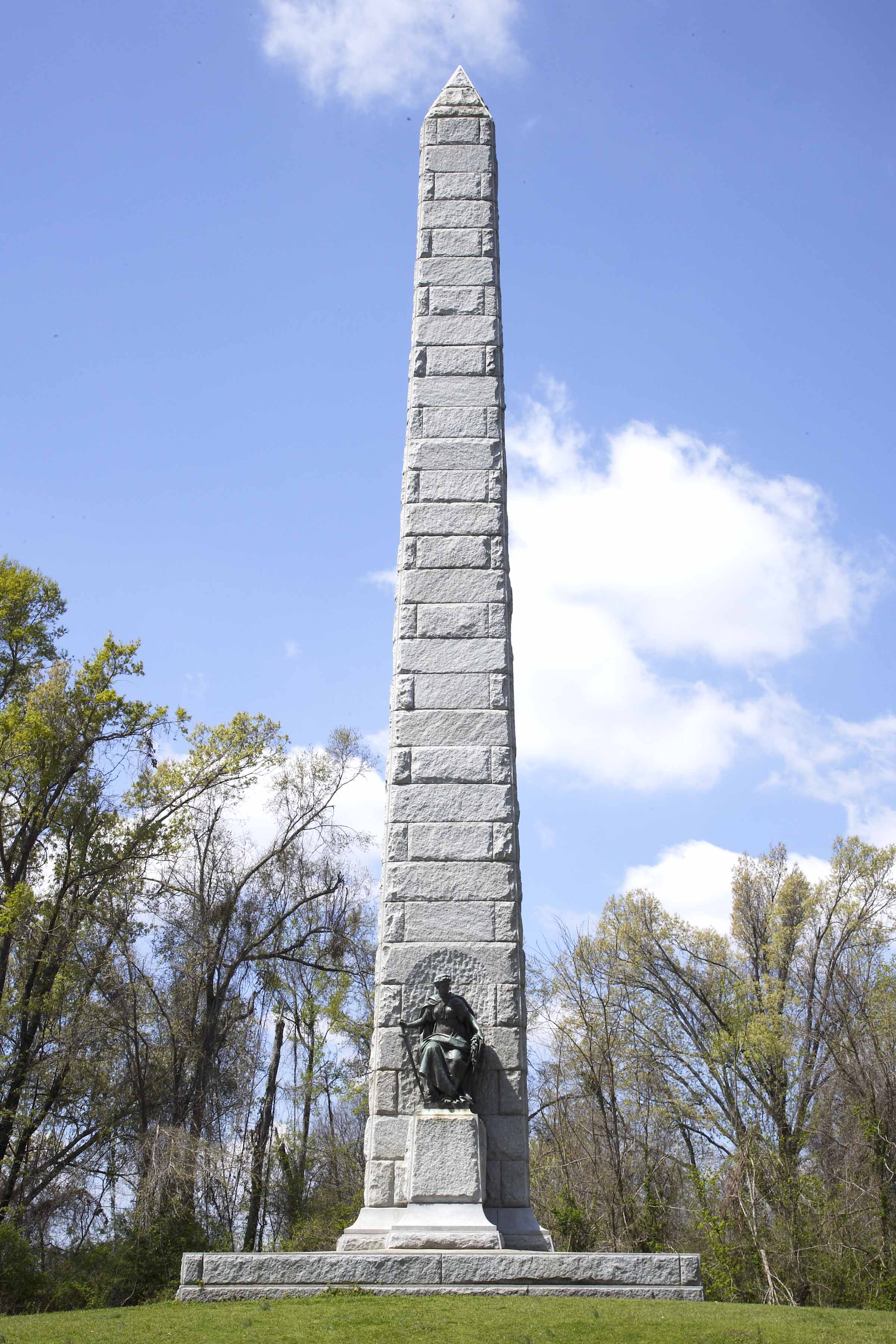 A tall white granite obelisk with bronze plaques on each side and in the center is bronze figure.