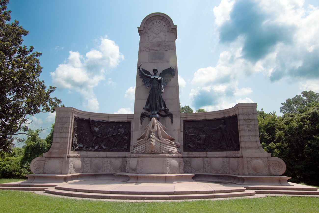 A large bronze statue of a woman with bronze relief images on left and right side of her.