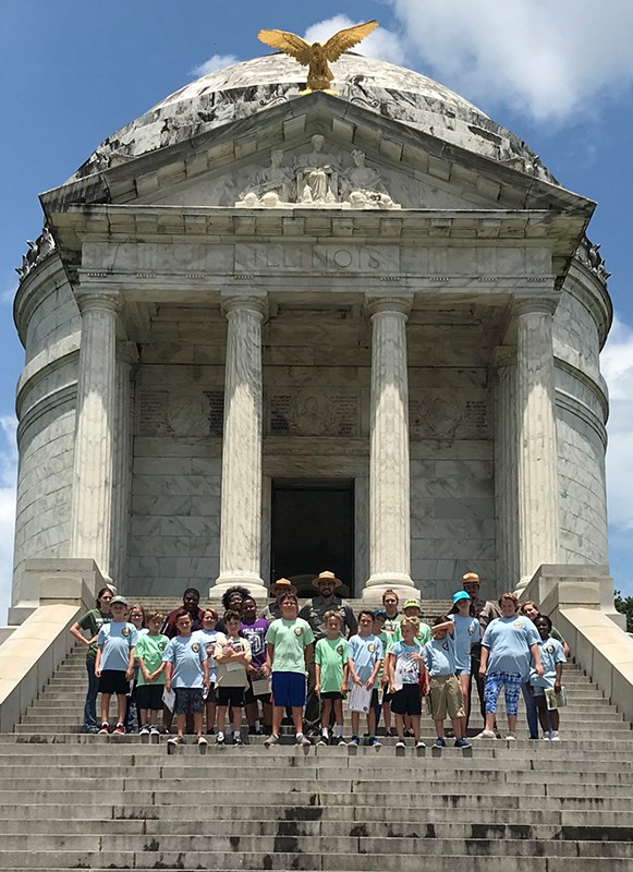 A group of park rangers and kids standing in front of a tall, rotund granite monument.