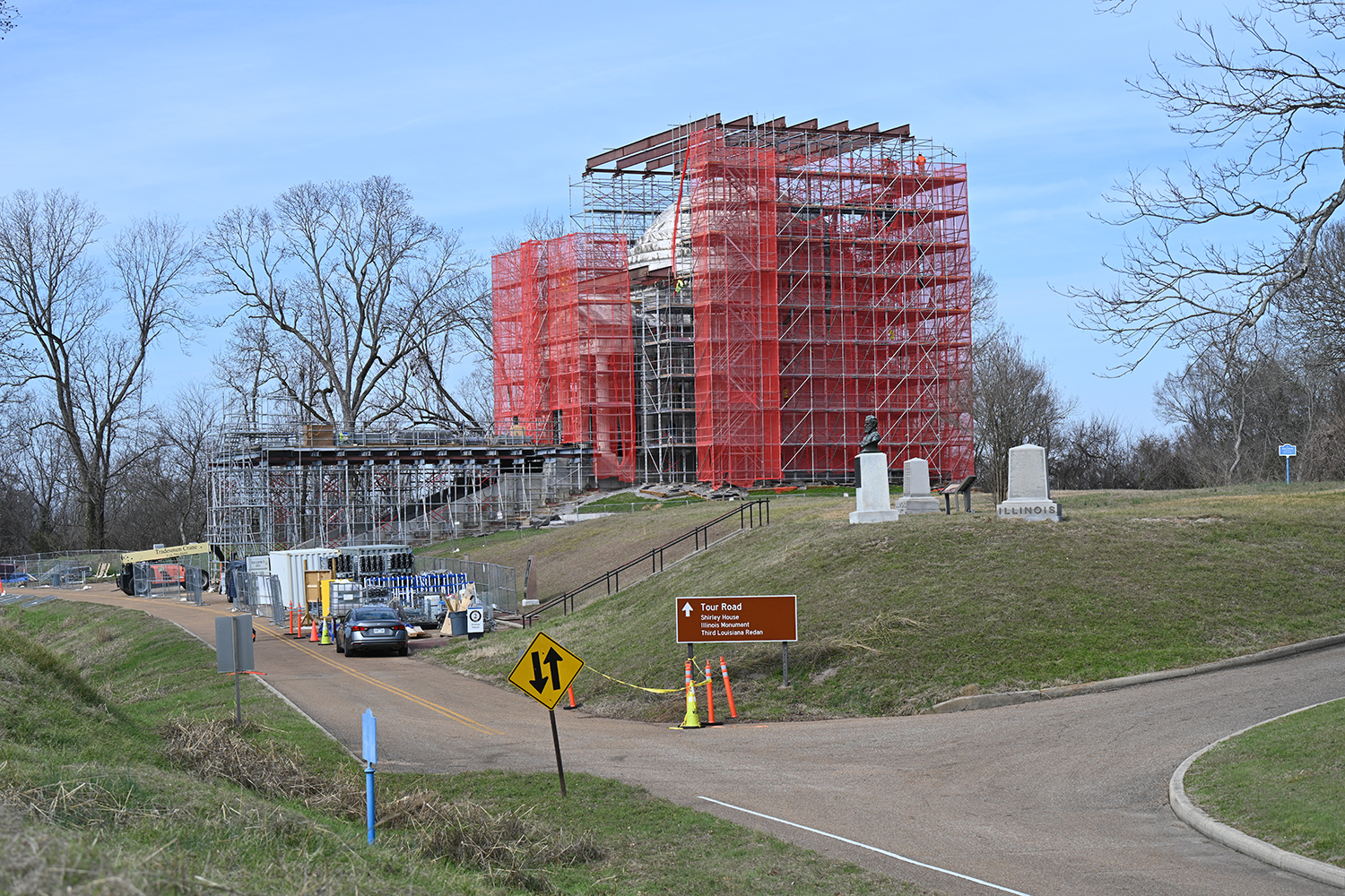 Large white stone structure on a hill above a road surrounded by scaffolding. Next to it is white house.