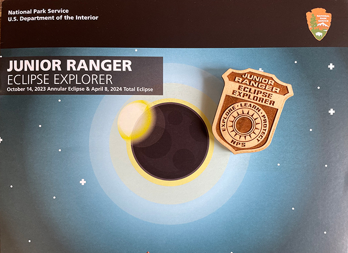Woode jr ranger badge with solar ecli[se written on it on top of a booklet with an artwork showing a solar eclipse over mountains.