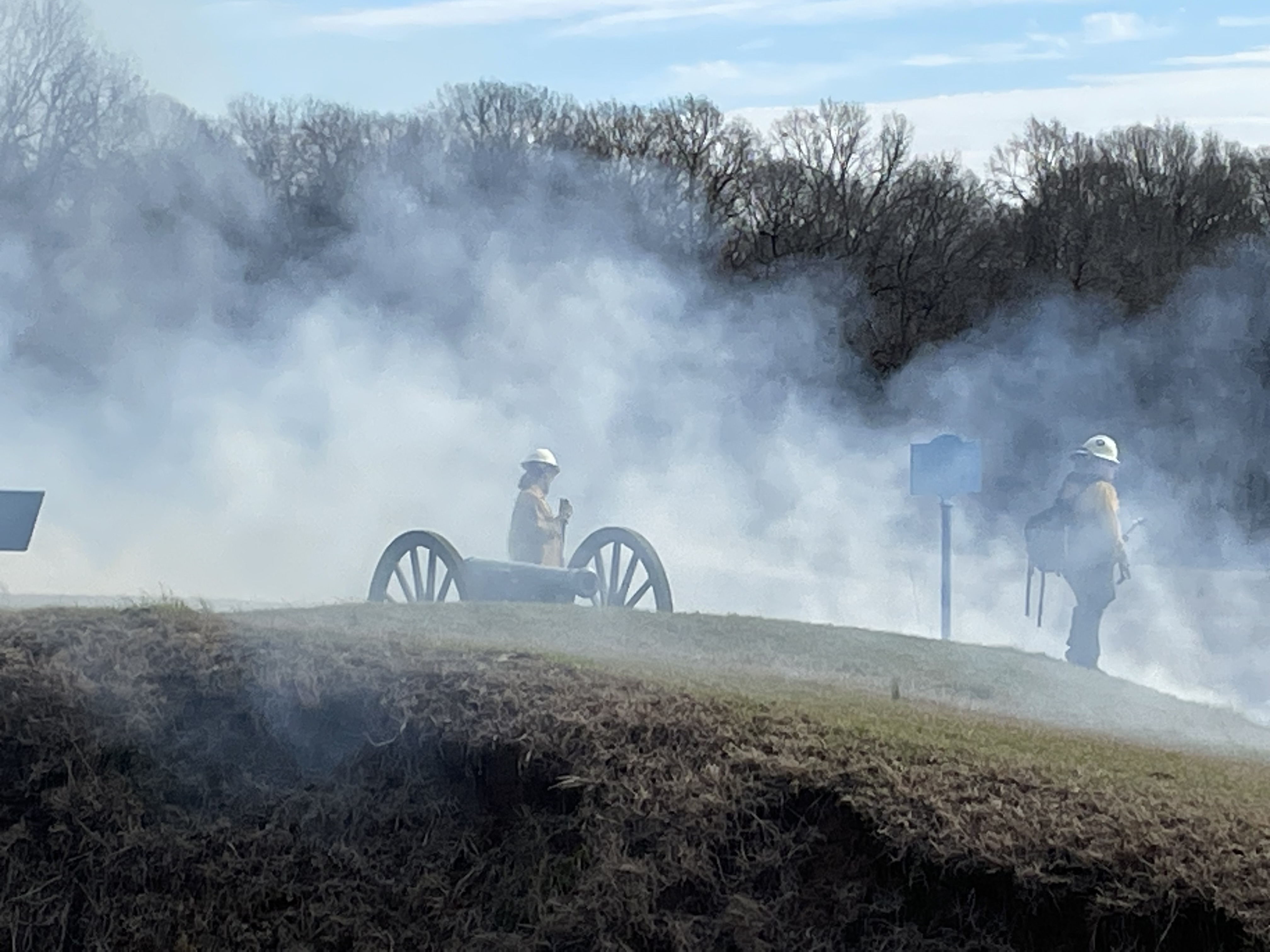 Small hill with a Civil War cannon on top with trees in backgrond. Two wildland firefighters are stand next to the cannon surrounded by wispy white smoke.