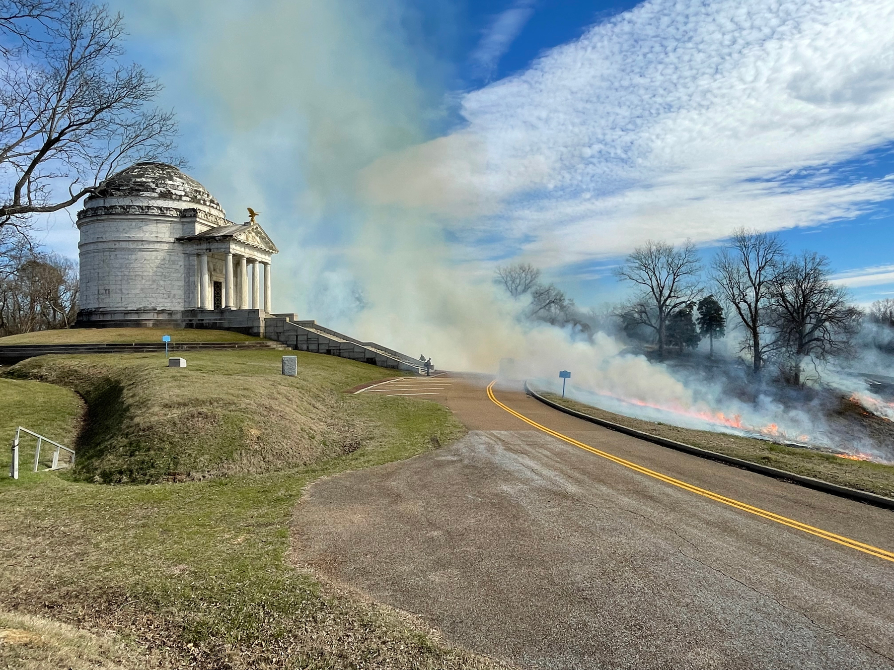 On the left is large white stone building on small hill with a dome and stairs leading up to it. In the center is road. On the right of the road firefighters mange a fire line with flames showing.