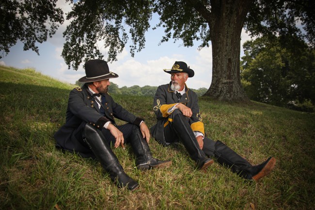 Full color image of two Civil War reenactors in the uniforms of a general sitting on a green grassy hill below a tree.
