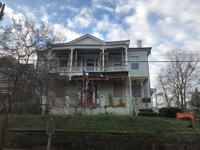 Building with scaffolding holding up to two level front porch. Orange cones on the right of the building block off a flight of stair leading up to the building. Blue sky with clouds above the building