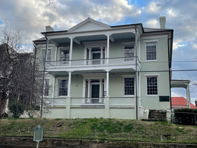 Light green two-story building with white trim. Sit op on raise are with a stone retaining wall and short black iron fence in front. Two story porch on front. Matching doorways on first and second level with columns framing doorways