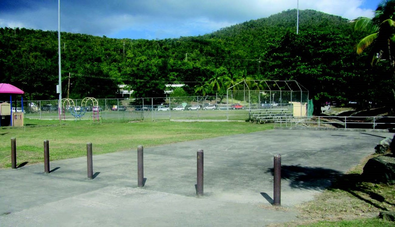 Concrete square with brown bollards in front and playground and fenced-in baseball field with green grass and tan dirt behind.
