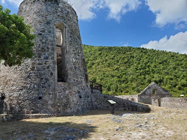 The grey and brown stonework of the windmill at Annaberg Plantation rises out of the rocky ground with the boiling house and animal mill in the background.