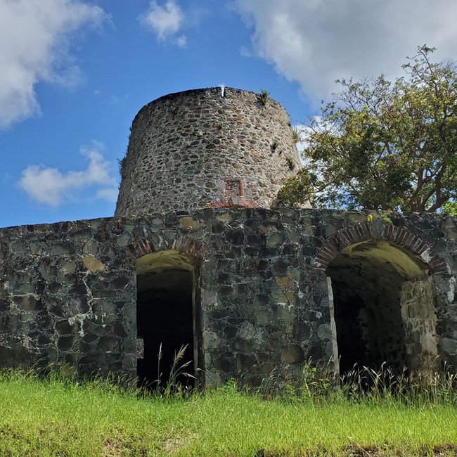 A stone cylindrical windmill ruin rises above two open masonry bays beneath, with blue skies behind.