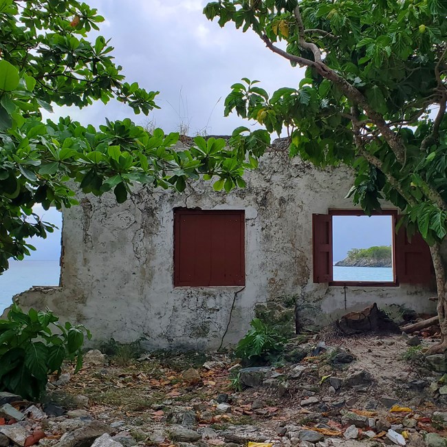 Green leafy trees frame a partially destroyed gray masonry building. One window with red shutters is open to the white sand beach and ocean view behind.