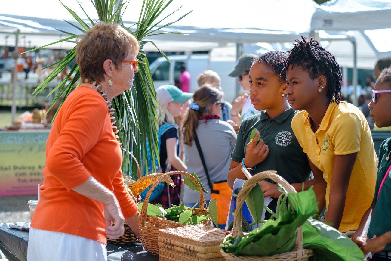 A woman in bright orange shares green plant leaves from baskets with three young students in polo shirts.