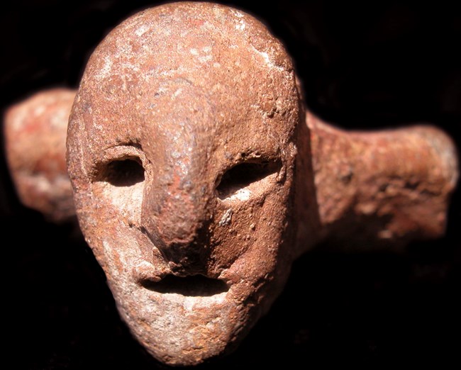 An image of a Zemi with a human face including eyes, a nose, and an reddish brown rock is pictured against a black backdrop.