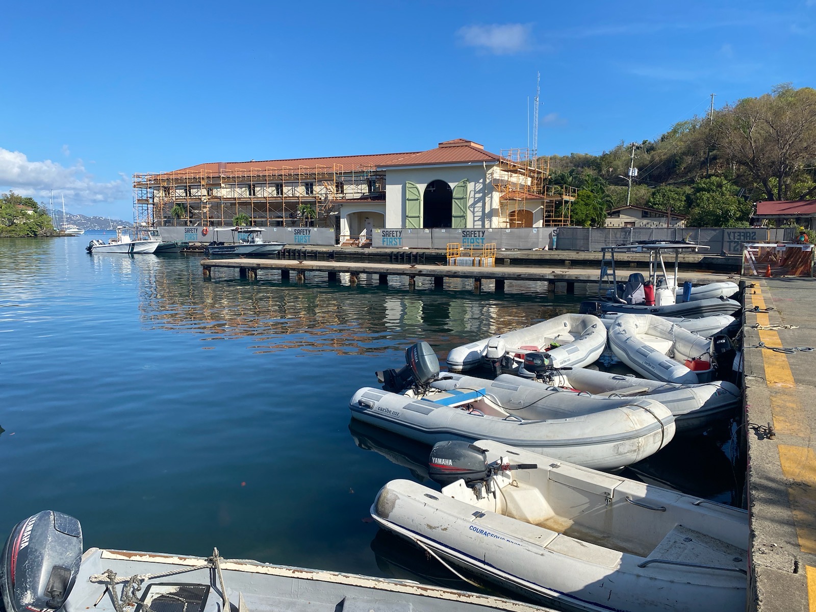 Nine small vessels are moored to the NPS bulkhead on the blue water of Virgin Islands National Park with copper roof and cream walls of the Cruz Bay Visitor Center in the background.