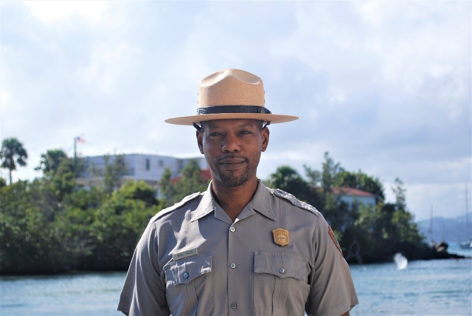 Man in NPS uniform stands in front of green, flowery bush with blue sky.