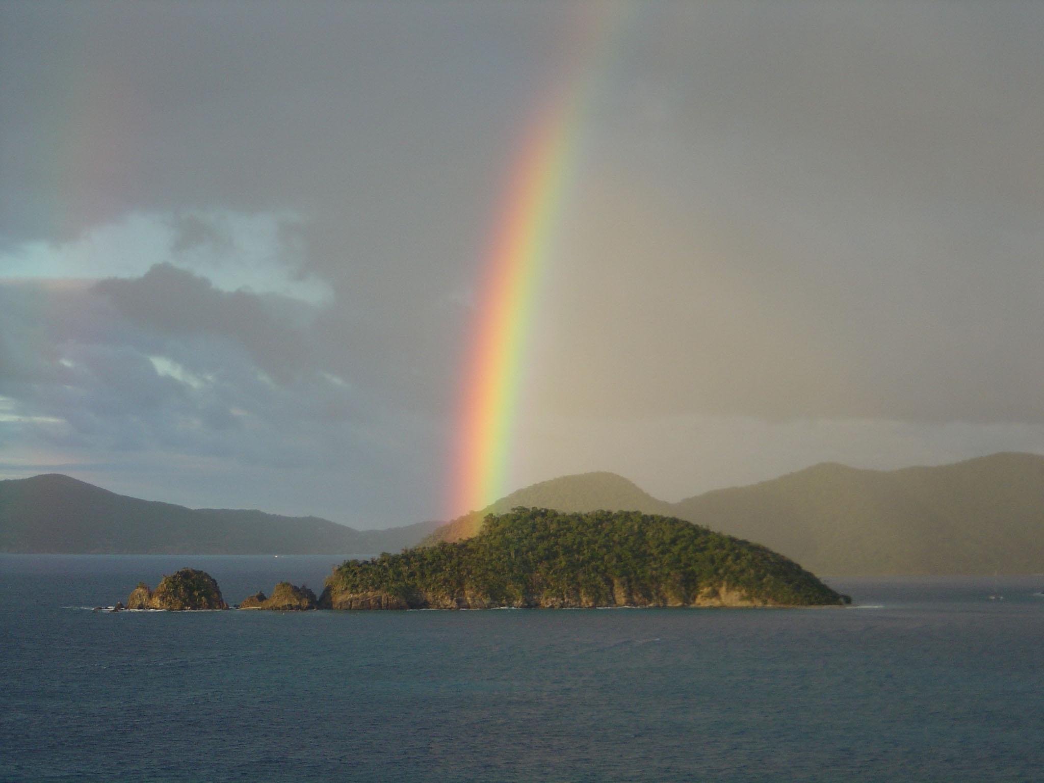 A rainbow stretches out over Whistling Cay, which protrudes from the blue waters of Virgin Islands National Park with its brown rocky coastline and lush green forested slopes.