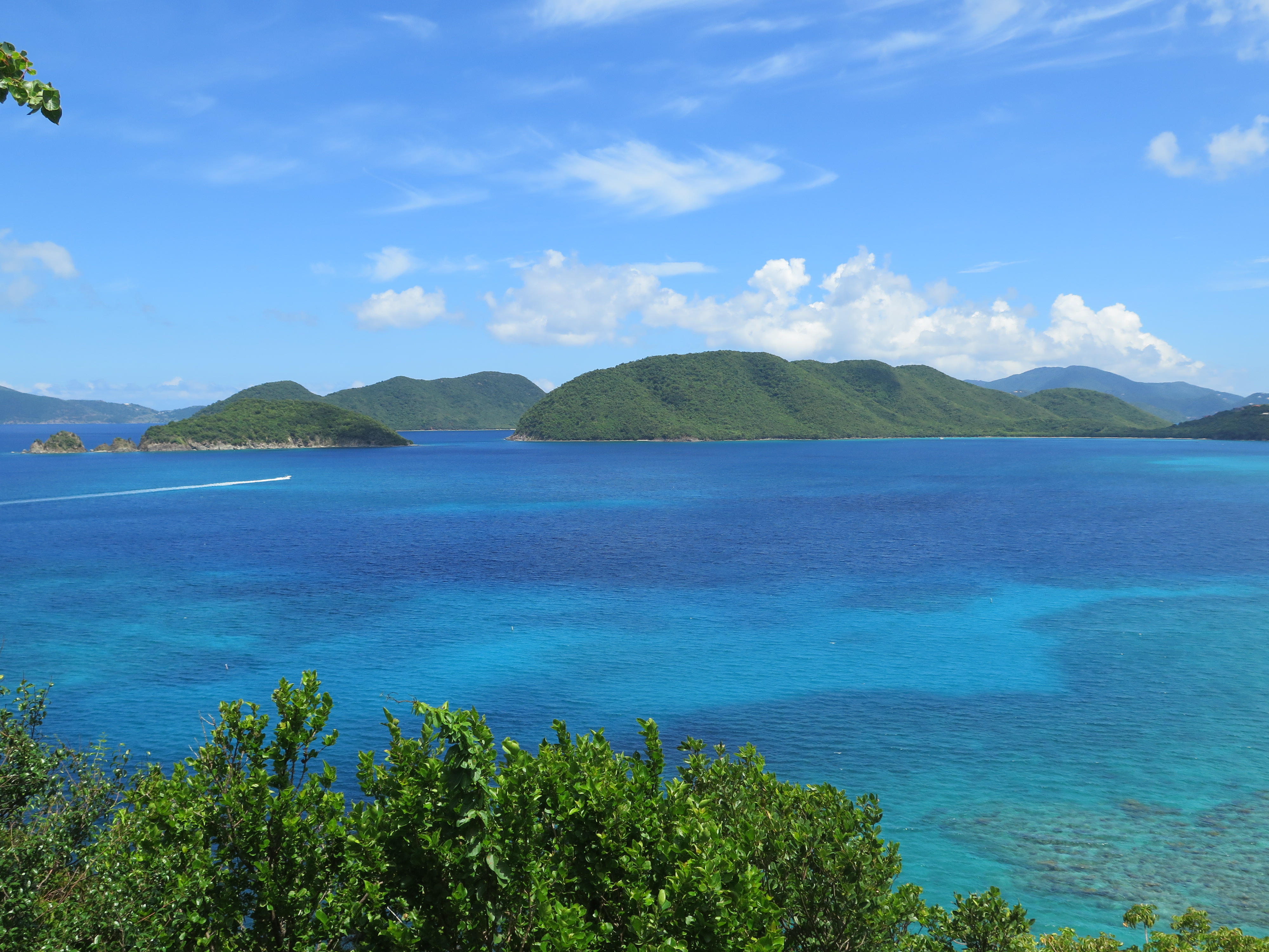Looking across the blue waters of Virgin Islands National Park, the forested green slopes of Whistling Cay, May Point, and Great Thatch Island stand tall beneath a blue sky dotted with white puffy clouds.