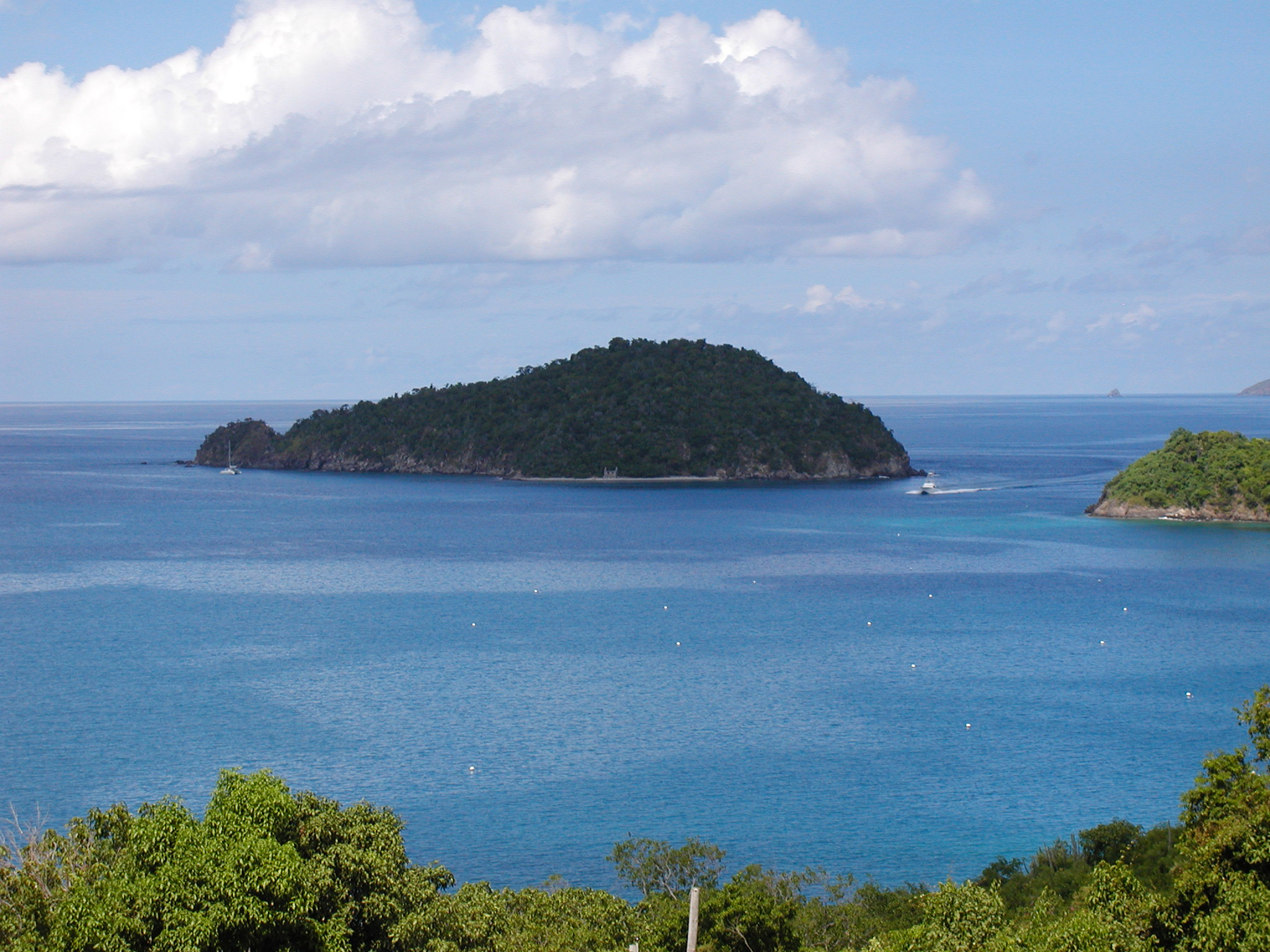 The grey rocky cliffs and forested green hillsides of Whistling Cay rise out of placid blue water beneath a partly cloudy sky.