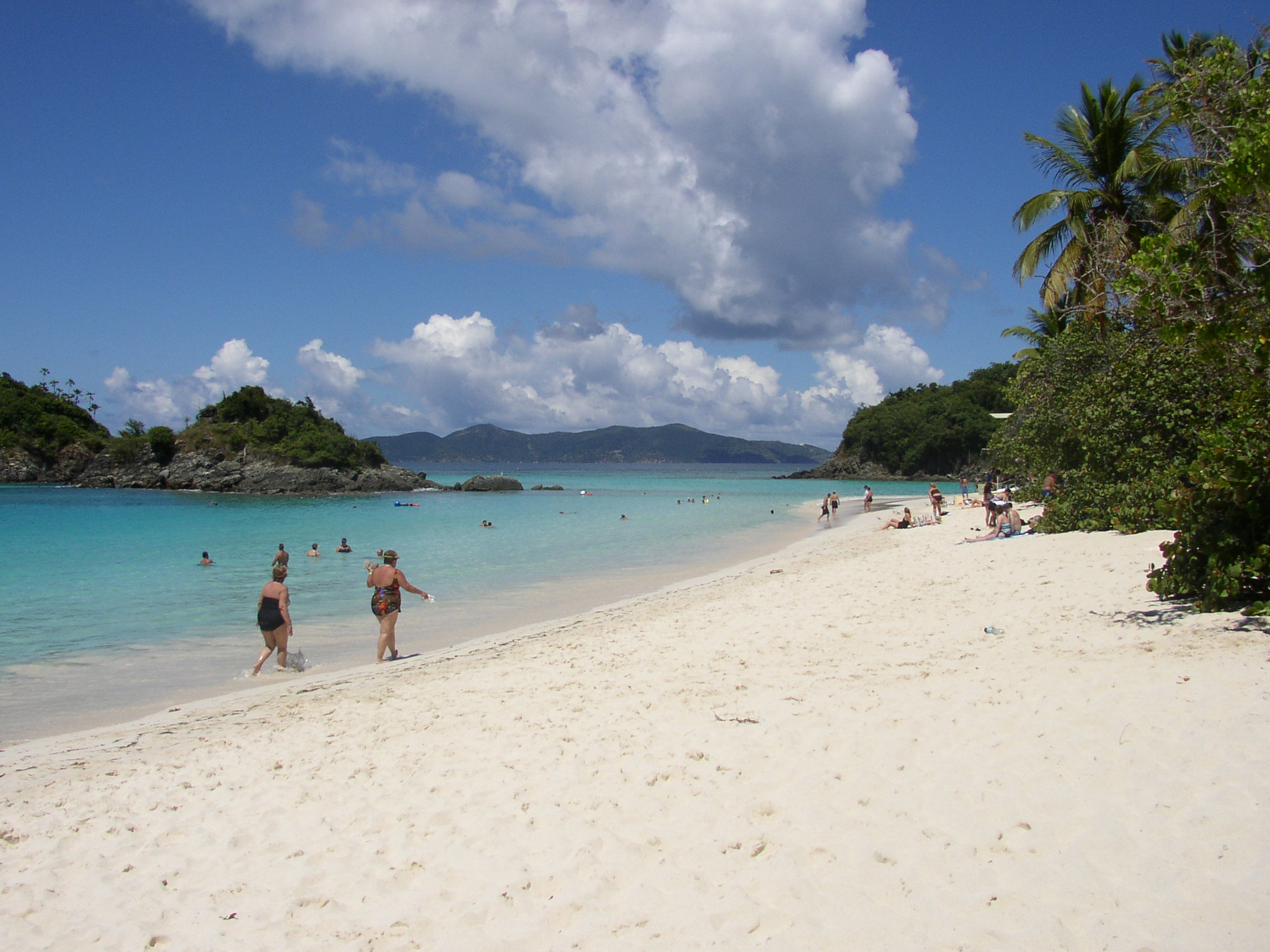 A group of visitors walk along the water's edge, wade, and relax on the white sand and blue water of Trunk Bay Beach.