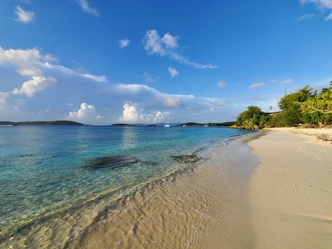 The white sand and crystal clear water of Salomon Bay are pictured under a blue sky streaked with soft white clouds.