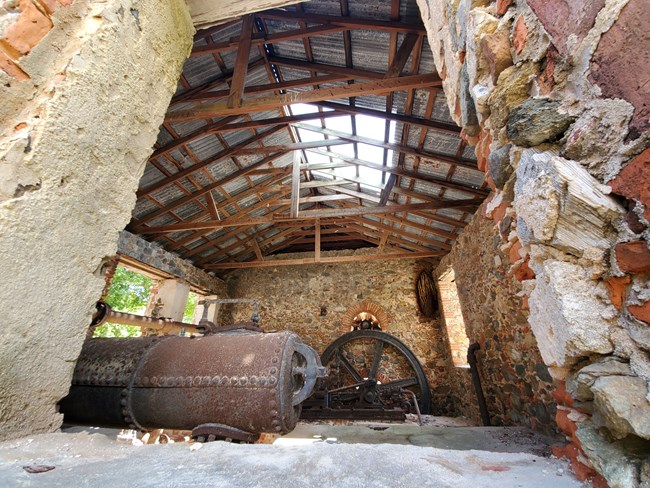 Reef Bay Sugar Factory's engine room is pictured through a window, revealing the rusty 19th century steam engine, Danish era ballast brick, field-stone, and coral construction, and reconstructed roof.