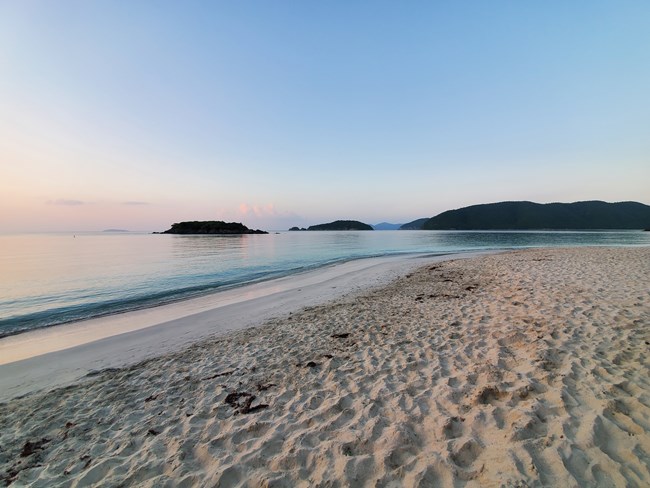 The glassy clear water of Cinnamon Bay are pictured from the white sandy shoreline as Cinnamon Cay, Whistling Cay, and Mary's Point appear to float on the horizon beneath a clear, blue sky.