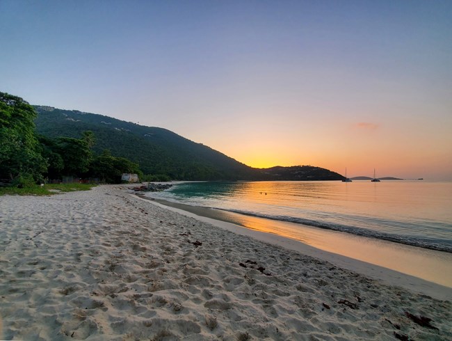 The white sand of Cinnamon Bay Beach gives way to placid ocean water as the last rays of the day's sun cast an orange yellow glow onto its surface from over the green hillsides of Virgin Islands National Park.