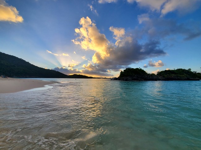 A view from Trunk Bay Beach depicting calm turquoise water lapping against a white sand beach with Trunk Cay in the background under a sun streaked blue sky with puffy white clouds.