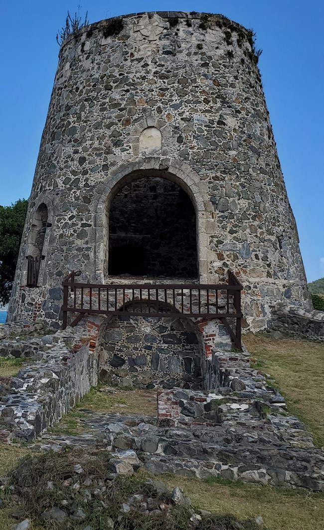 Under a clear blue sky, the Annaberg Plantaion windmill, with its grey stone, brick, and coral construction, rises out of the green grass.