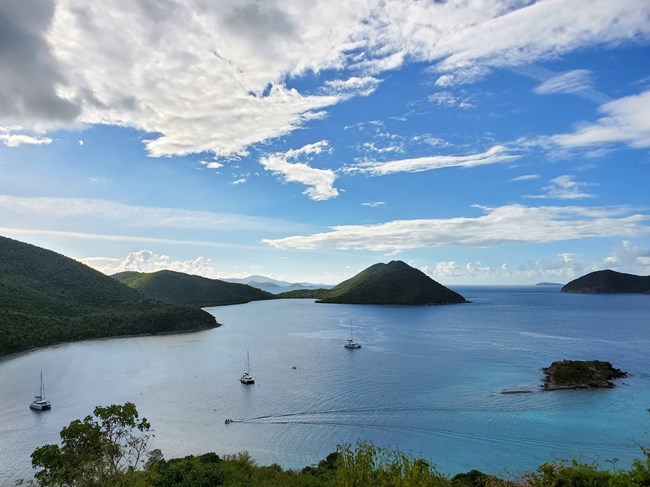 The blue waters of Leinster Bay and Mary's Creek are pictured from above with St. John's rolling green hills stretching into the distance and a blue sky with puffy white clouds.