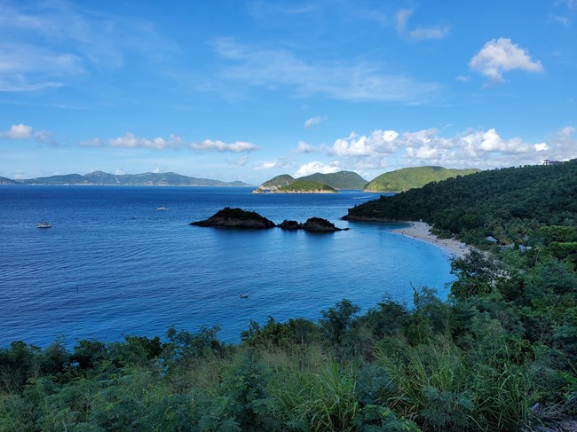 Beneath a blue sky dotted with white clouds, the beautiful blue waters of Trunk Bay extend from the beach's white sandy shoreline with Trunk Cay's three iconic peaks jutting out of the ocean just offshore of the beach.