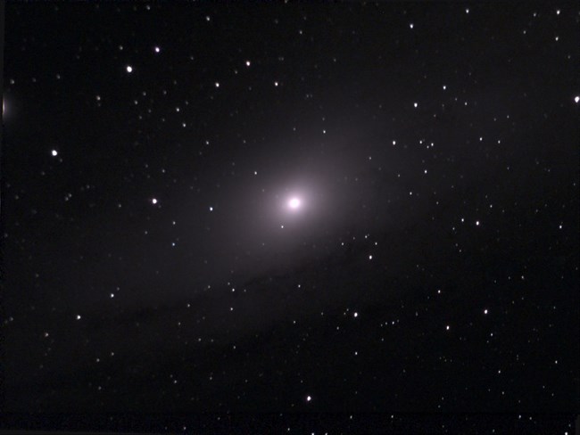 A group of billions of stars called the Andromeda Galaxy appears as a hazy cloud against the black backdrop of interstellar space. The individual stars in the foreground are part of the Milky Way Galaxy.