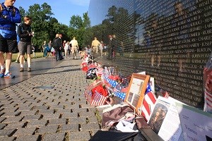 Visitors are on a sidewalk looking at a black reflective wall and thousands of mementos left by family and friends.