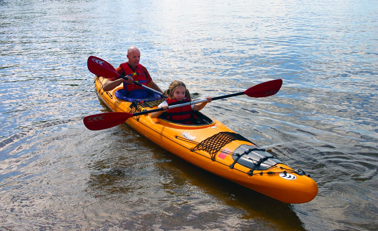 A school-aged girl and her father paddle a two-seated tandem kayak by the shore of a scenic blue lake.