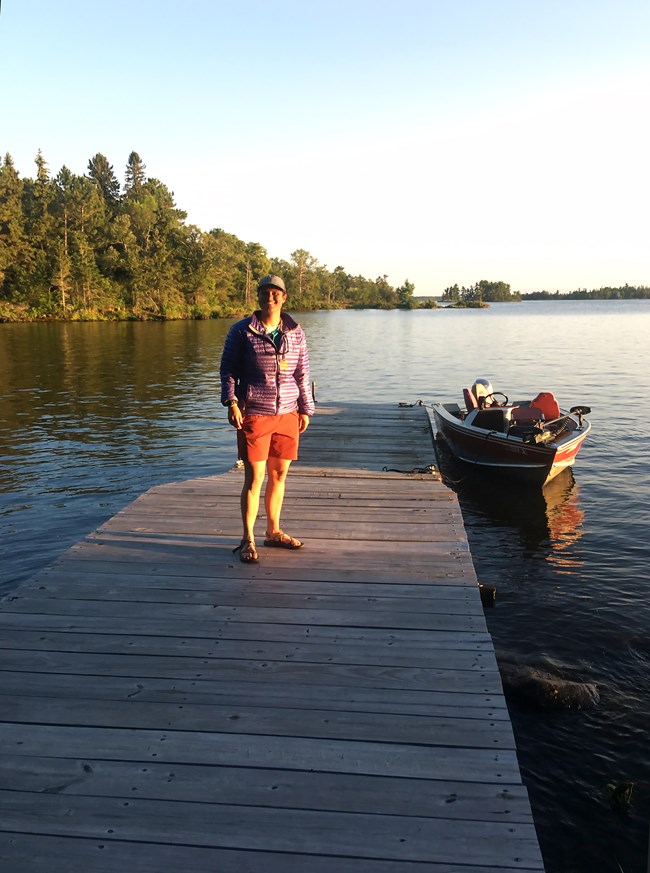 A woman in a purple jacket stands on a dock on the shore of a scenic lake. A red boat is tied to the dock next to her.