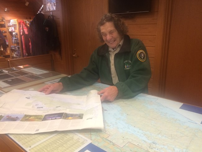 A volunteer in a tan uniform smiles and holds out a map from behind the front desk of a visitor center.