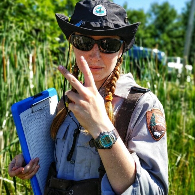 ield technician in sun hat and sun glasses stands in marsh with a clipboard, holding up two fingers in the sign for peace.