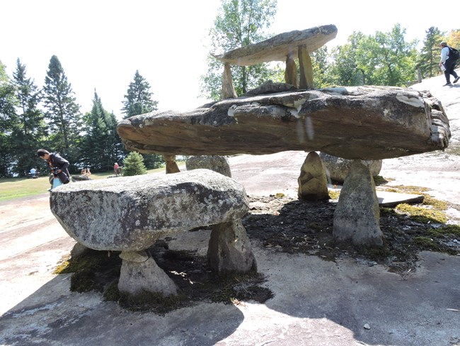 A two-layered table made from a large, stone slab balanced atop several small, pointed rocks sits upon a large, stone outcropping. Visitors wander in the background against a grassy lawn and scenic lakeshore.