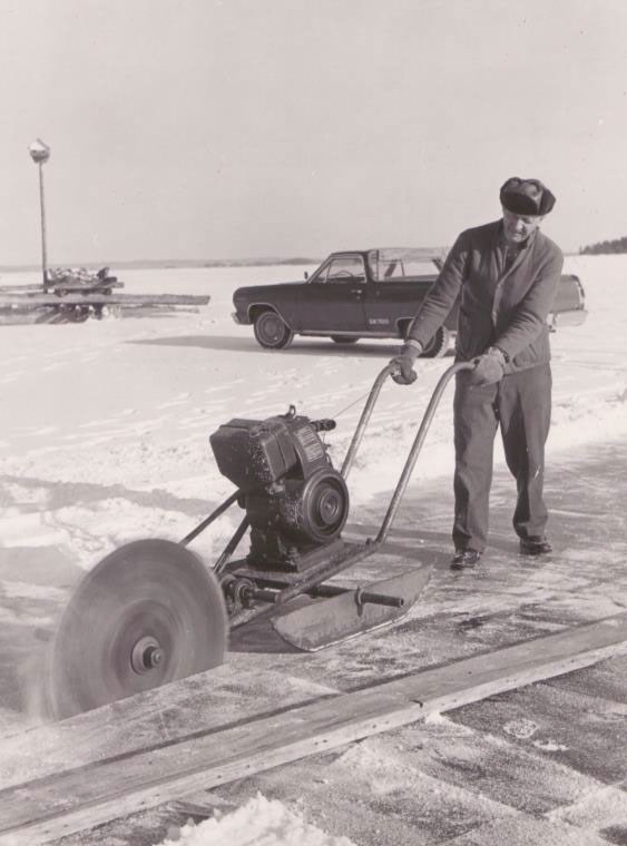 A historic photo of a man using a large saw to cut ice blocks from the frozen lake surface