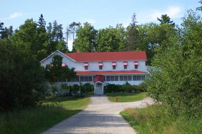 A two-story building, white siding, red roof and awnings, with large screened in porch along the front.  Surrounded by trees with a road leading up to the front and center of the building.