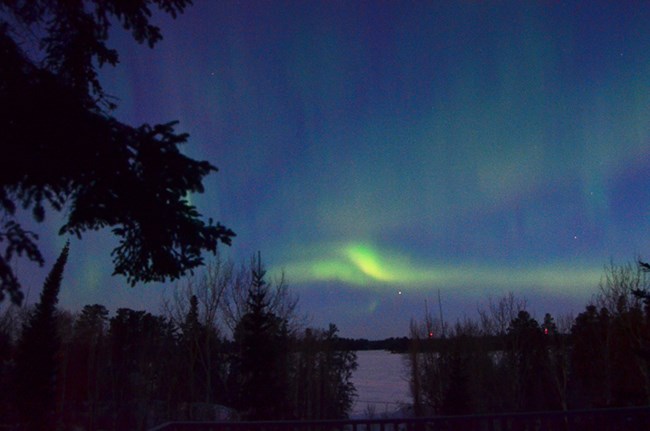 Several faint green curtains of light glow against dark skies above a frozen lake shore.