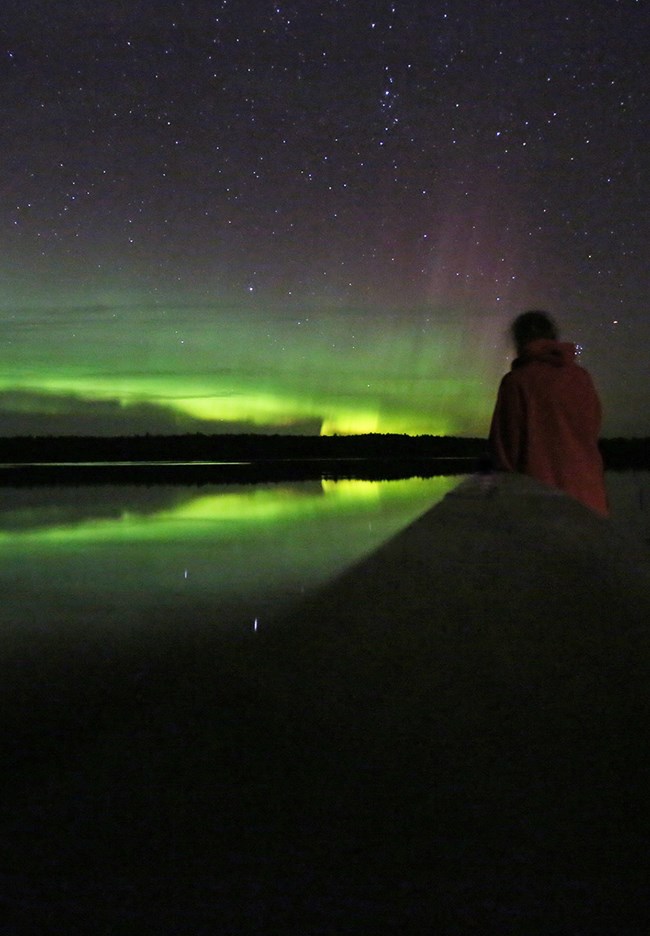 A silhouetted figure stands on a dock and watches a bright green curtain of light shining above a tree-lined horizon.