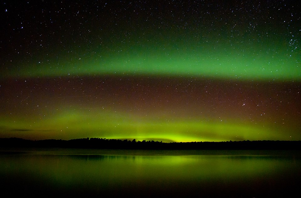 Two bright green curtains of light shine against a dark, starry sky over a tree-lined horizon and a scenic lake.