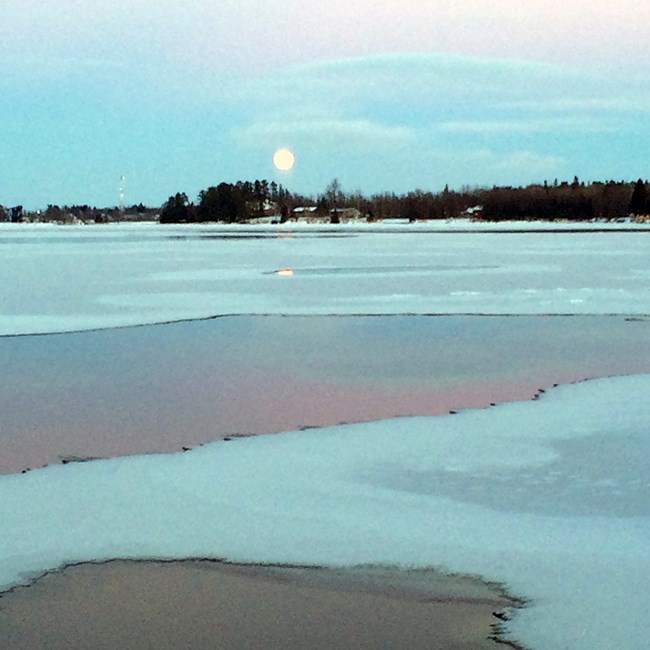 A full moon rises over the silhouette of a dark, tree-lined island on the shores of a scenic lake at dusk.