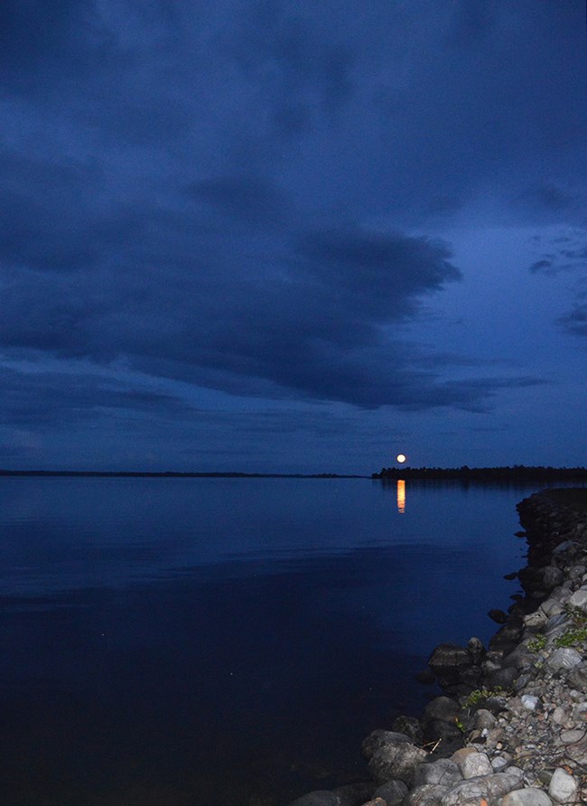 A full moon rises over the silhouette of a dark, tree-lined island on the shores of a scenic lake at dusk.