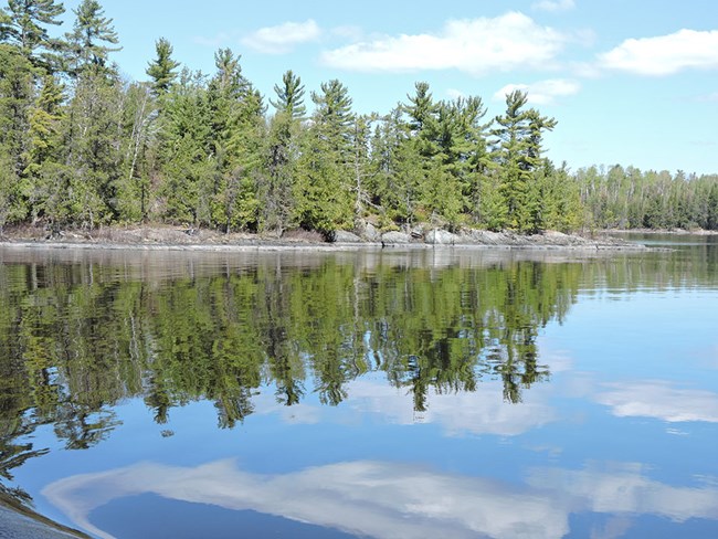 A reflection of trees, rocks, and blue sky on the waters of Rainy Lake