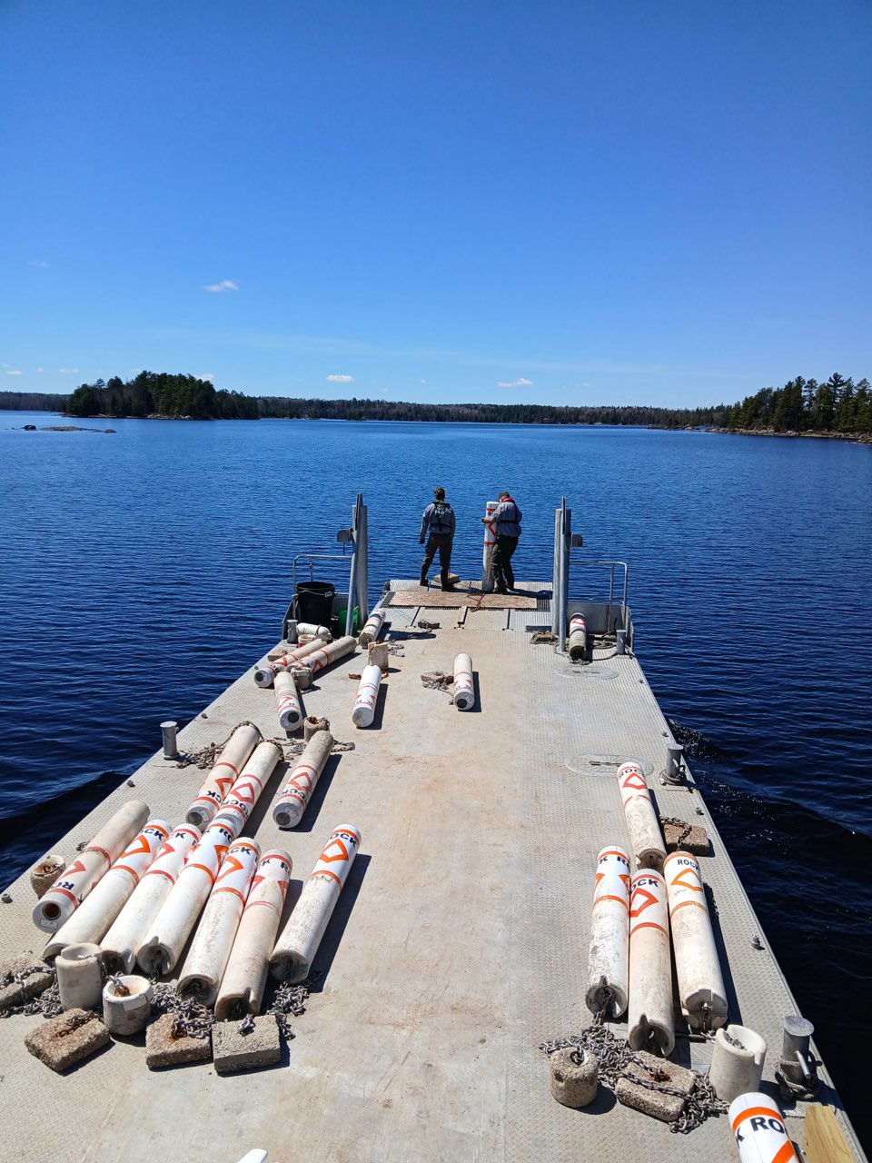 A long, grey barge boat is on the blue waters of Rainy Lake and there are blue skies overhead. On the deck are dozens of white rock hazard buoy markers and two staff people stand at the end ready to place one of the markers in the water.