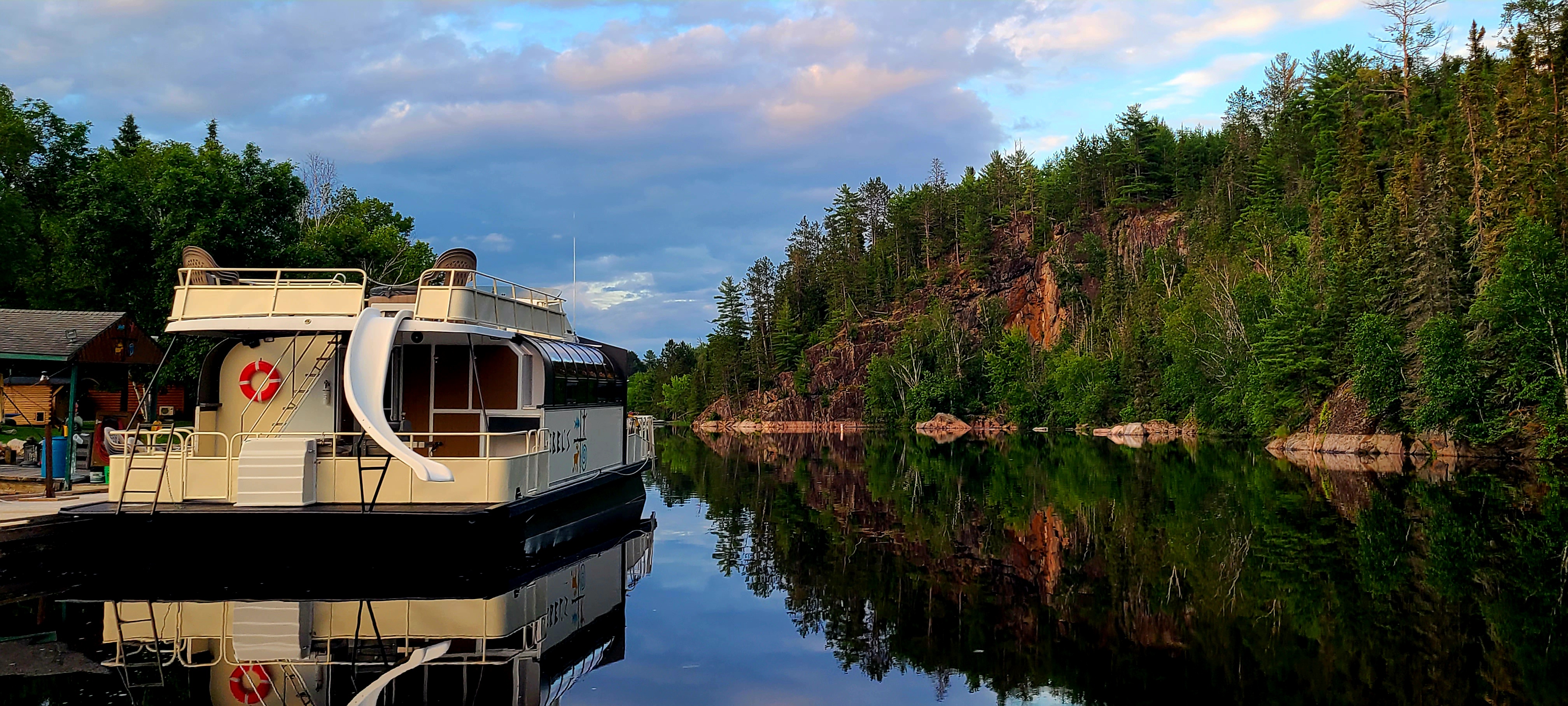 A houseboat shown from the backside that's on the water with a slide coming down from the upper deck. In the background are rocky cliffs, trees, and partially clouded skies, which are reflected of the still water.